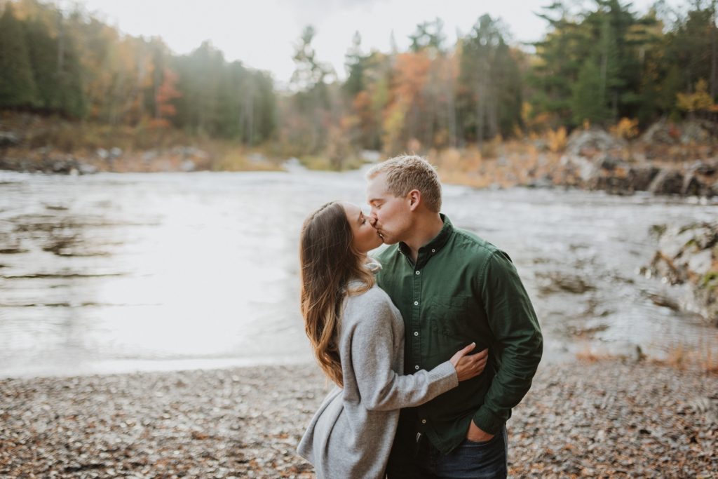 Couple kissing in front of river and woods. 